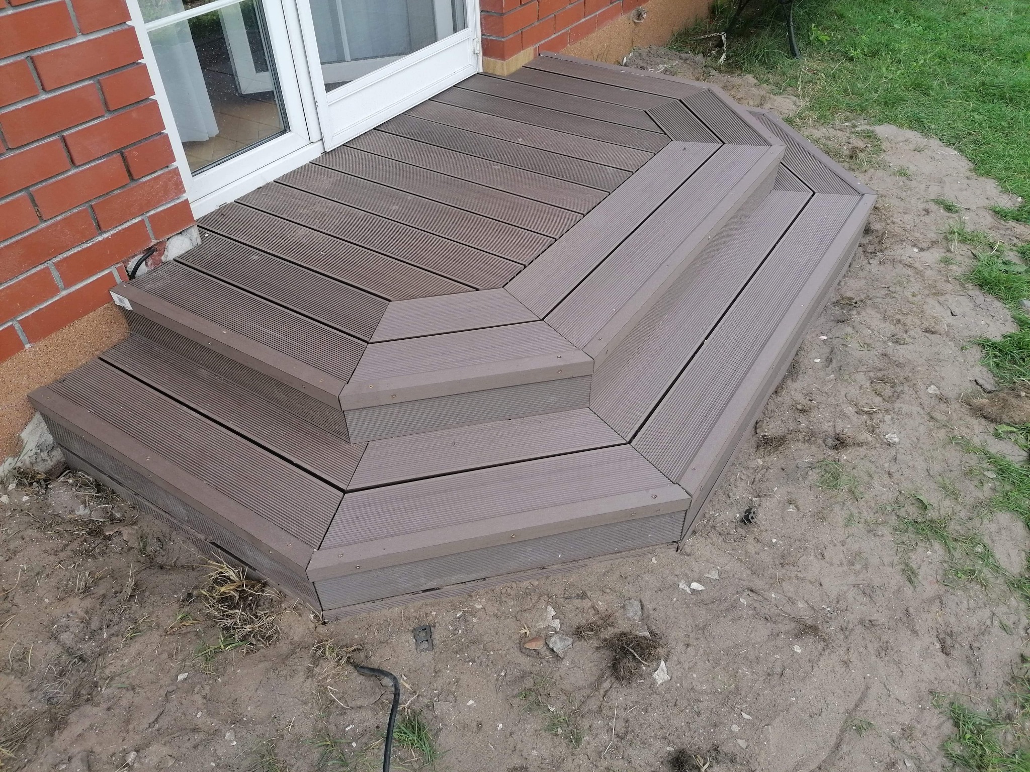 Nowoczesny taras z kompozytu w odcieniach brązu, z trzema stopniami, przylegający do ściany budynku z czerwonej cegły i białych drzwi balkonowych, otoczony ziemią i fragmentami trawy.