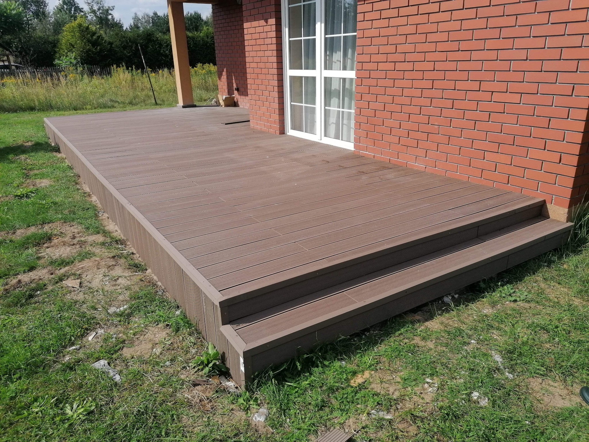 Nowoczesny taras z kompozytu w kolorze brązowym, przylegający do ceglanego domu z białymi drzwiami balkonowymi, widok na trawnik i zieleń w tle.