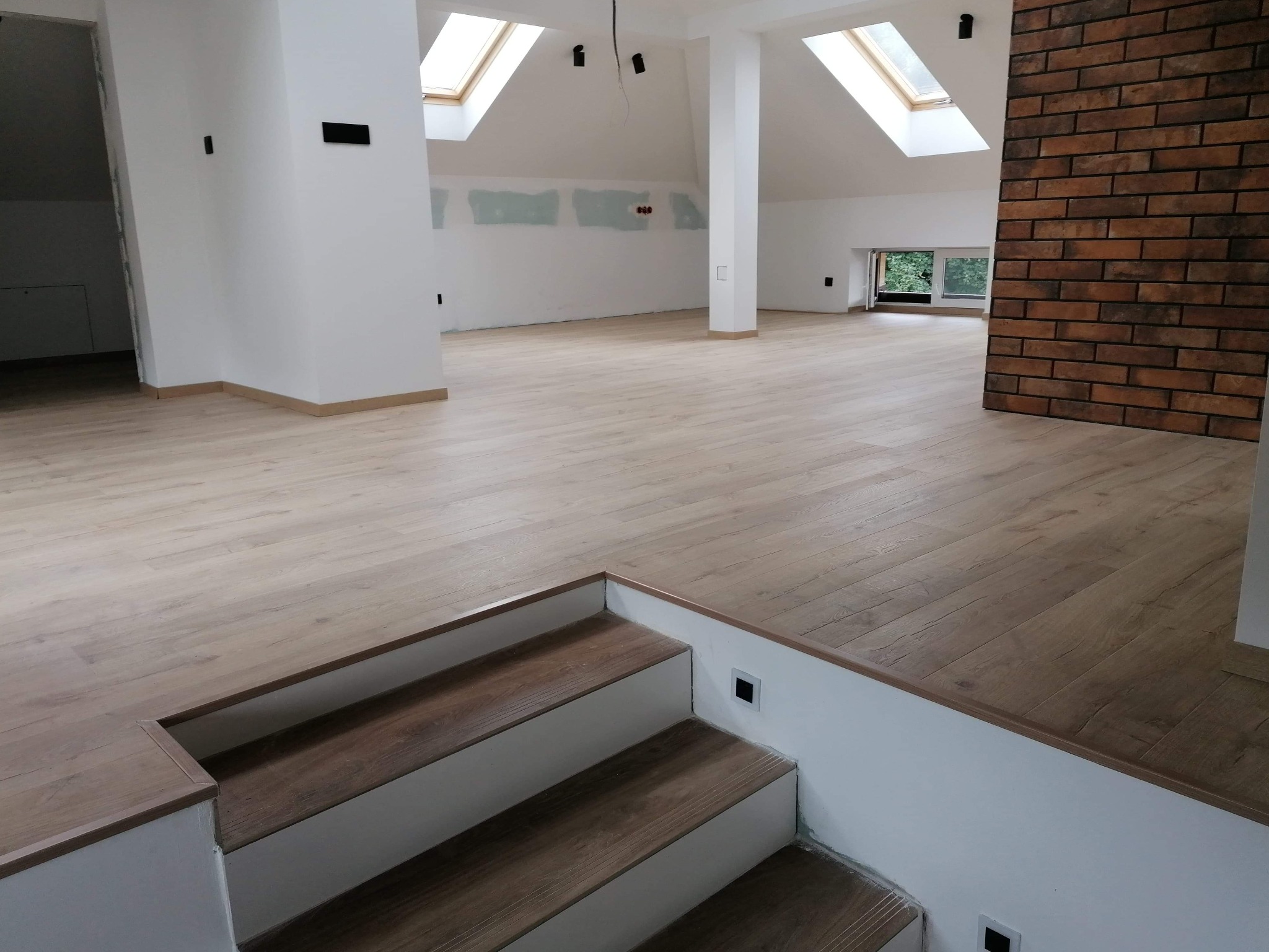 Jasna podłoga z paneli w przestronnym, jasnym poddaszu z oknami dachowymi i schodami z podświetleniem, ściana z cegły w tle.
