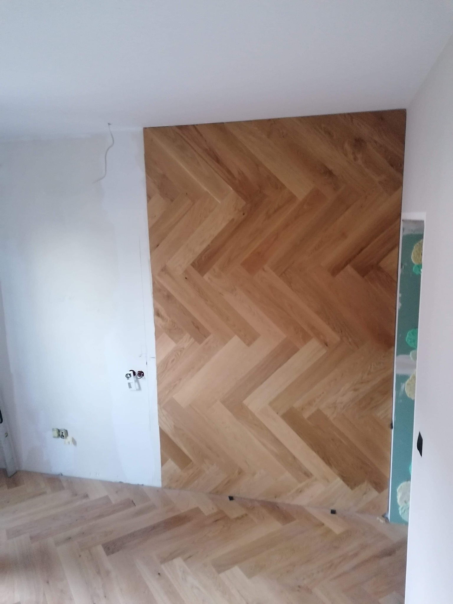 Fragment ściany i podłogi z ułożonym parkietem w jodełkę, widoczne niedokończone wykończenie ściany z instalacją elektryczną.