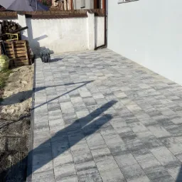 Nowo ułożona szara kostka brukowa na podwórku, widoczny cień osoby, biała ściana budynku w tle, paleta z drewnem i zwinięty wąż ogrodowy.