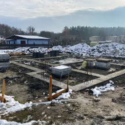 Fundamenty domu jednorodzinnego w zimowej scenerii, z widocznymi zbrojeniami, bloczkami i drewnianymi szalunkami, częściowo pokryte śniegiem, w tle zabudowa jednorodzinna i las.