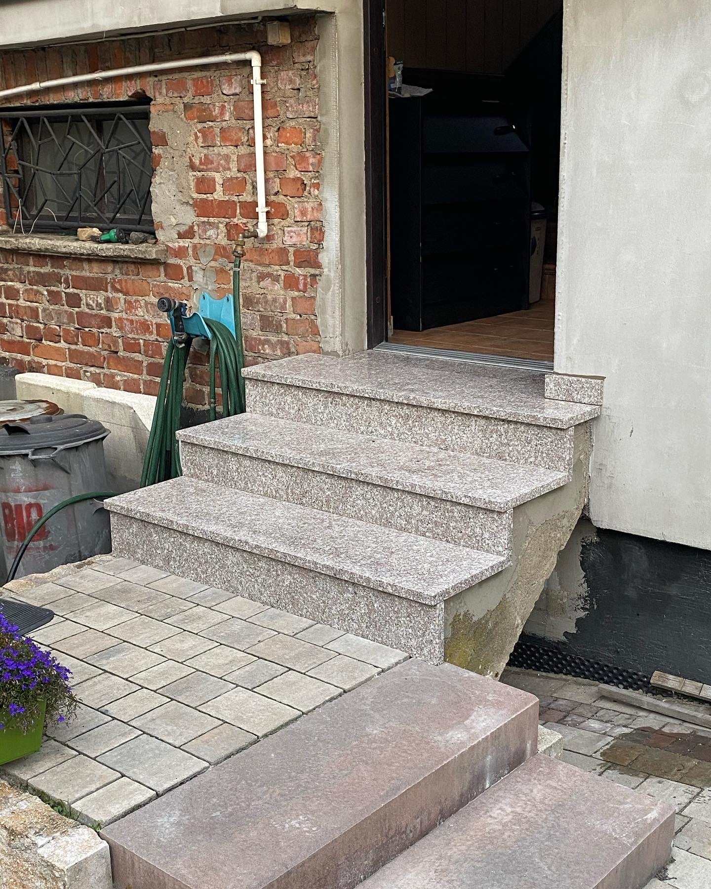 Granitowe schody wejściowe do budynku z ceglaną ścianą po lewej stronie, na której wisi wąż ogrodowy, obok stoi kosz na śmieci, a przed schodami ułożona kostka brukowa.