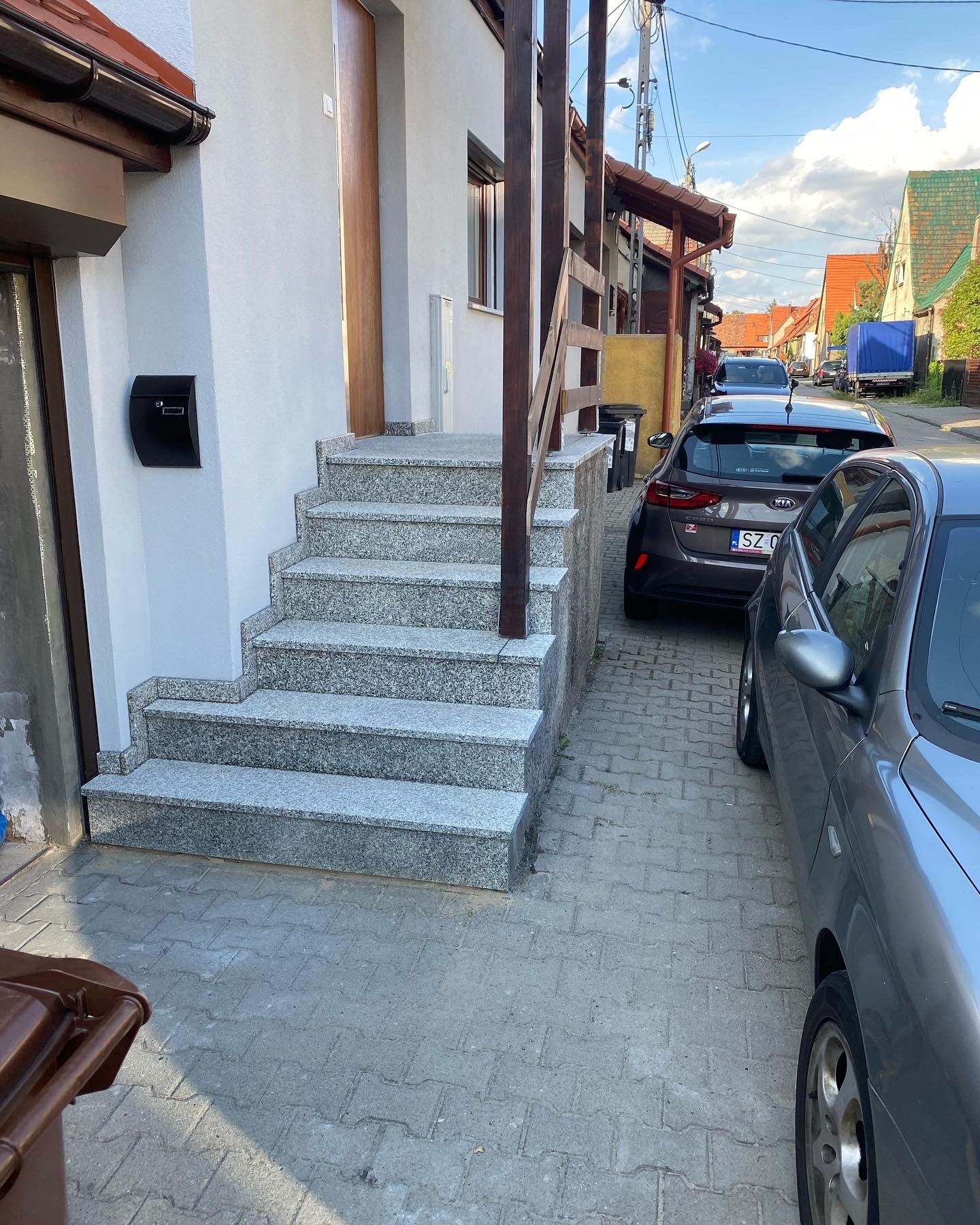 Granitowe schody wejściowe do domu z drewnianą balustradą, skrzynka na listy na ścianie, widok na ulicę z zaparkowanymi samochodami.