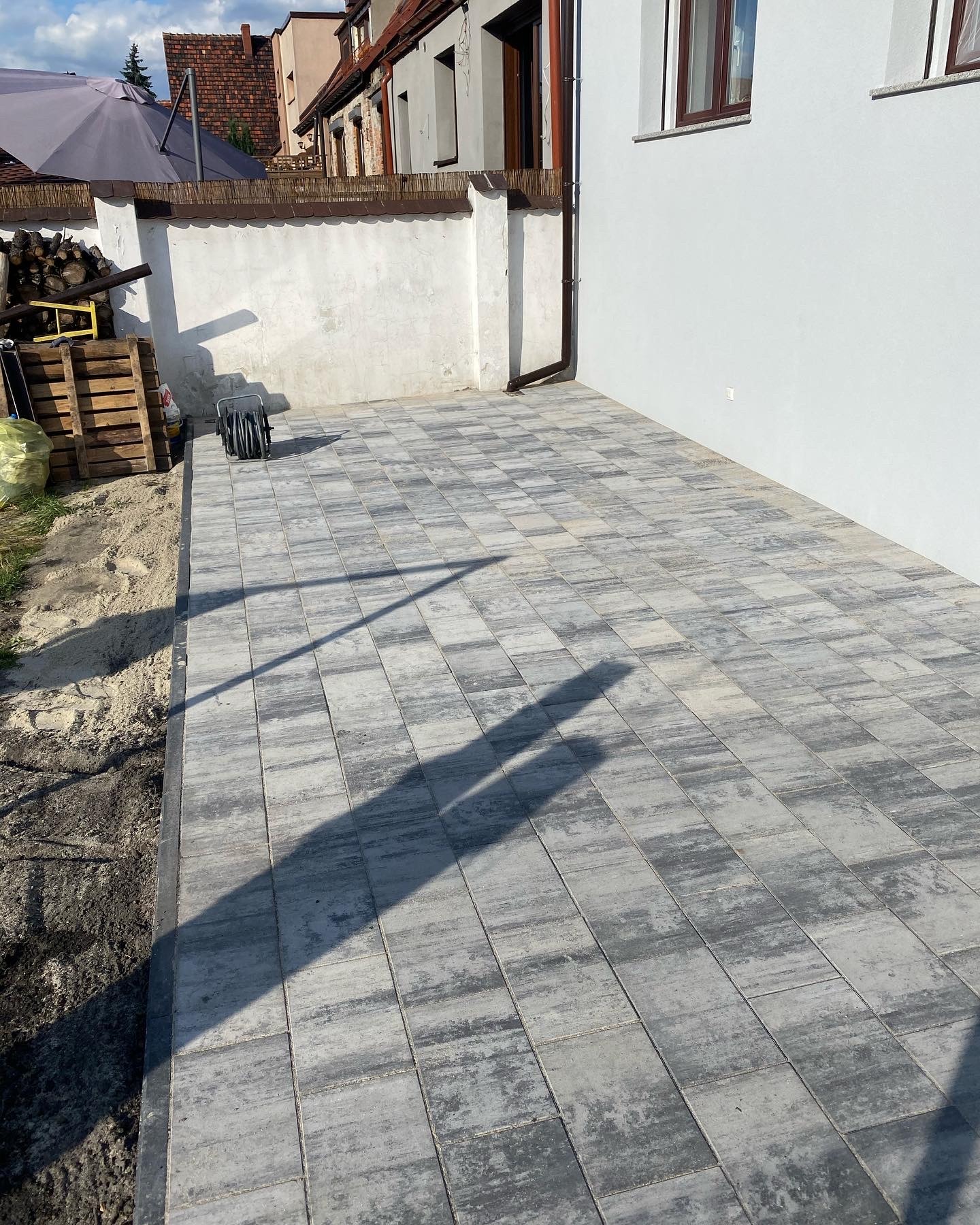 Nowo ułożona szara kostka brukowa na podwórku, widoczny cień osoby, biała ściana budynku w tle, paleta z drewnem i zwinięty wąż ogrodowy.