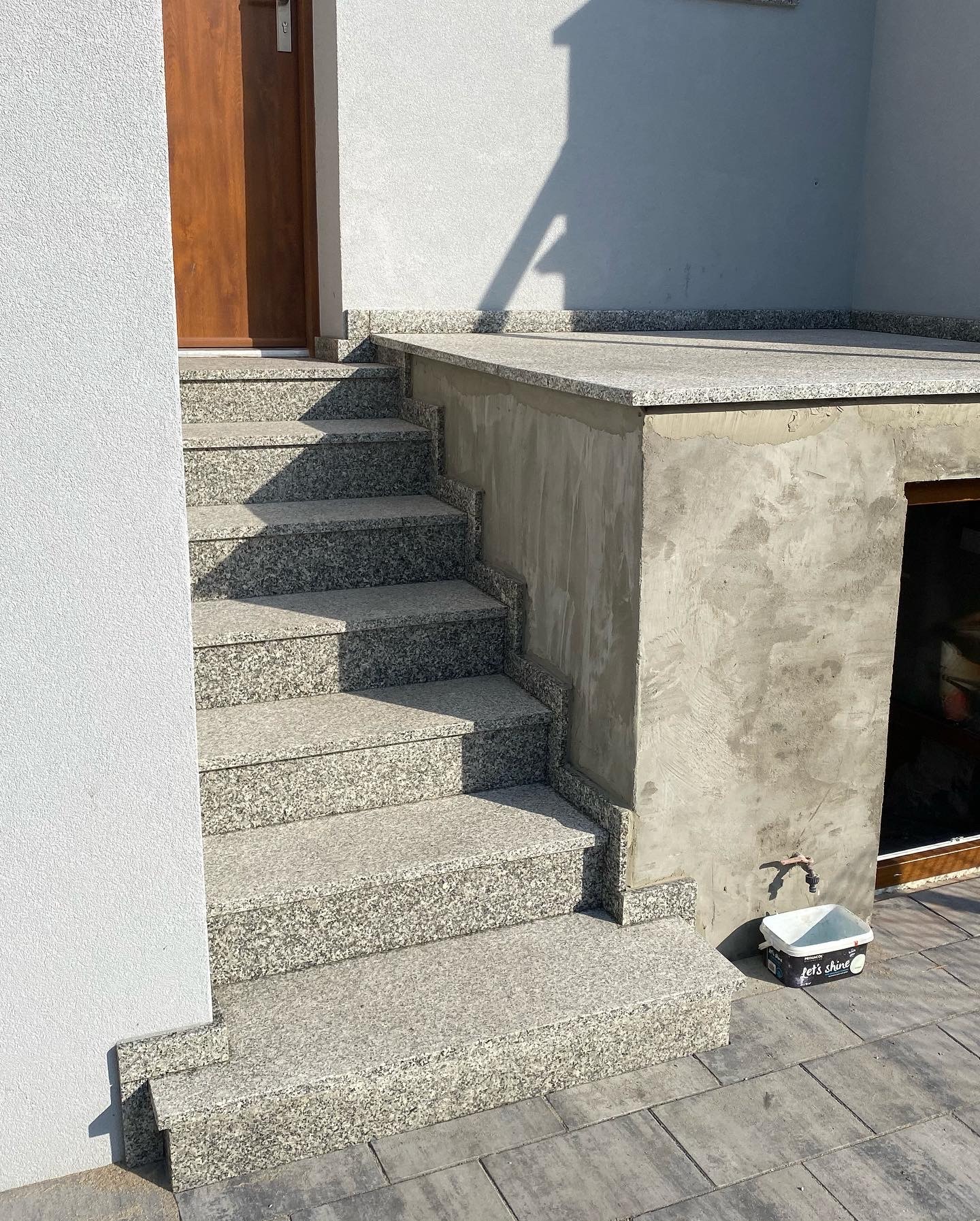 Schody zewnętrzne wyłożone płytami z jasnego granitu, prowadzące do drzwi wejściowych; obok ściana z surowego betonu z kranem i pojemnikiem na farbę.