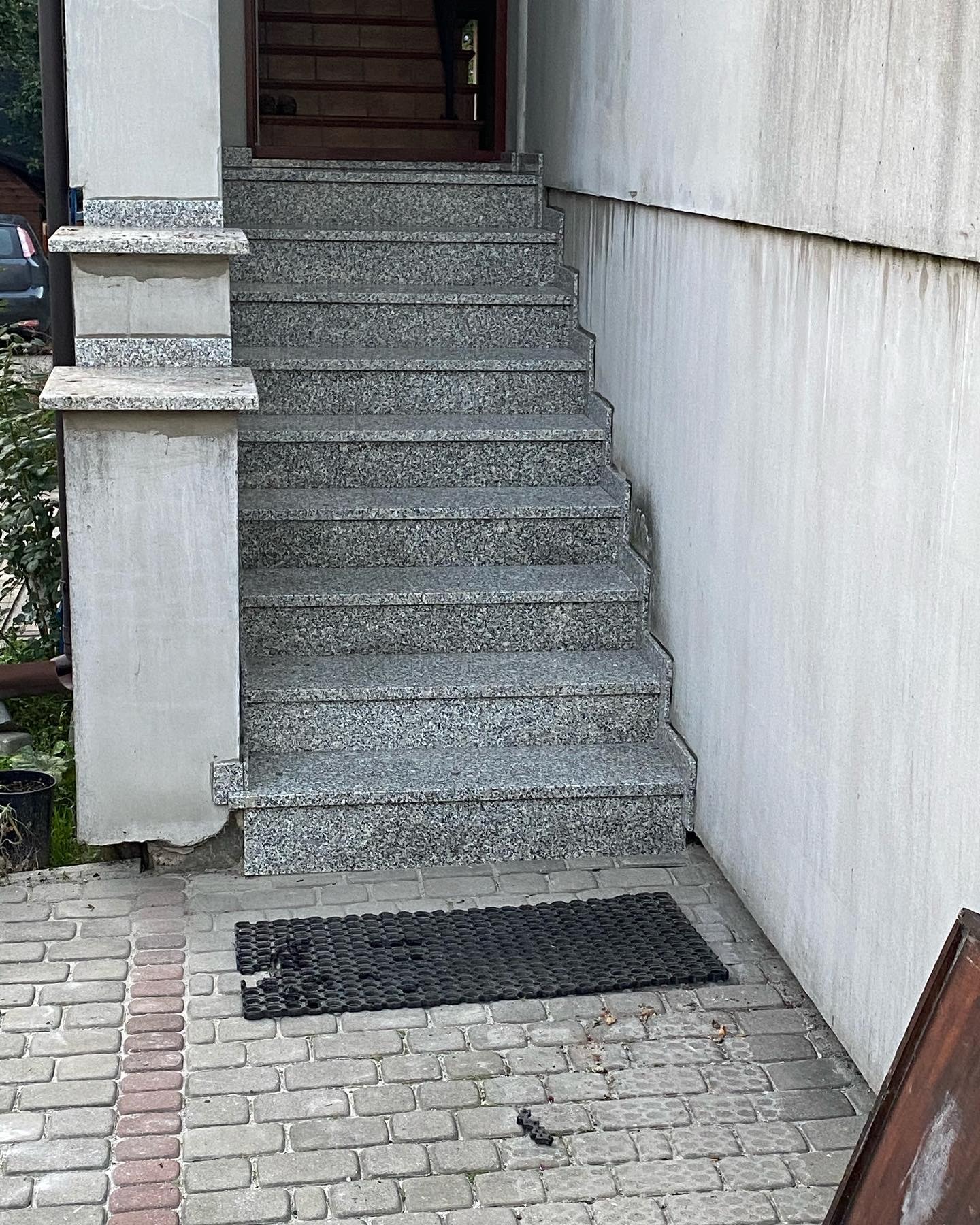Granitowe schody zewnętrzne prowadzące do wejścia budynku, z widoczną kostką brukową i gumową wycieraczką przed drzwiami, ściana budynku z widocznymi zaciekami.