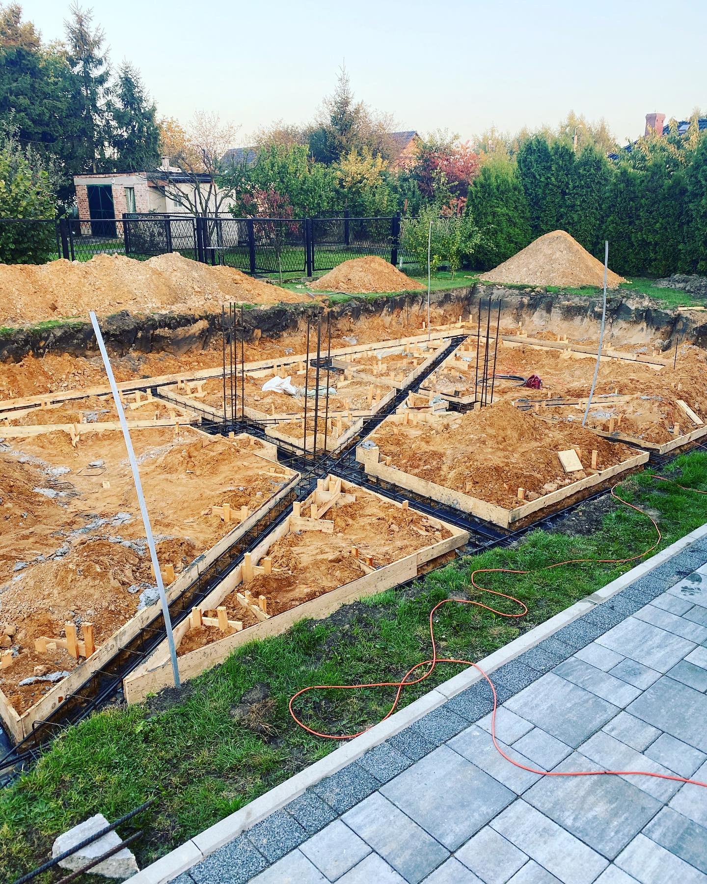 Przygotowanie fundamentów pod budynek: drewniane szalunki zbrojeniowe, wykop pod ziemią, hałdy ziemi w tle, widok z góry na plac budowy.