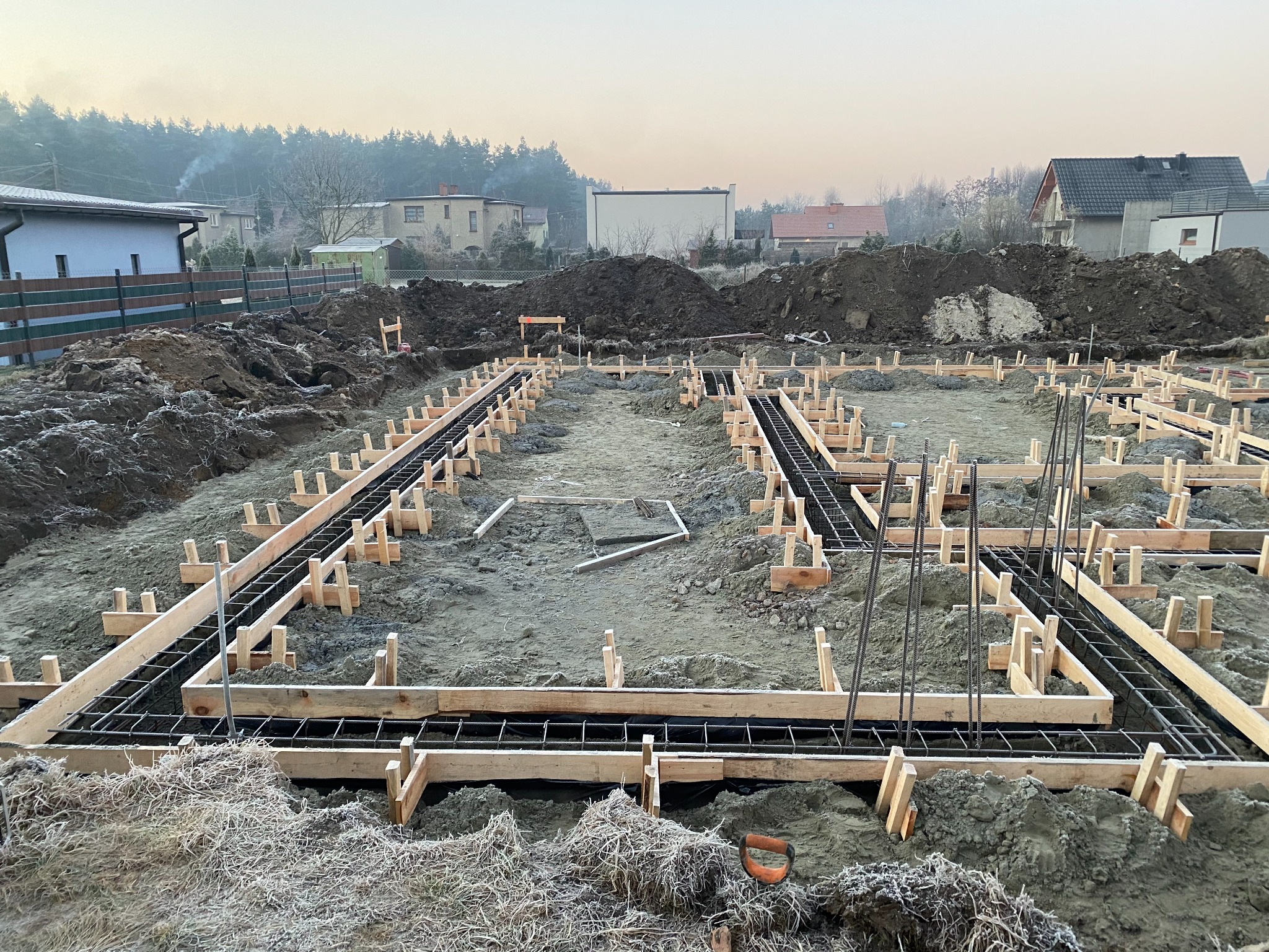 Przygotowanie fundamentów pod budowę domu: drewniane szalunki i stalowe zbrojenie na placu budowy, w tle zabudowa jednorodzinna i drzewa, poranek z lekkim mrozem.