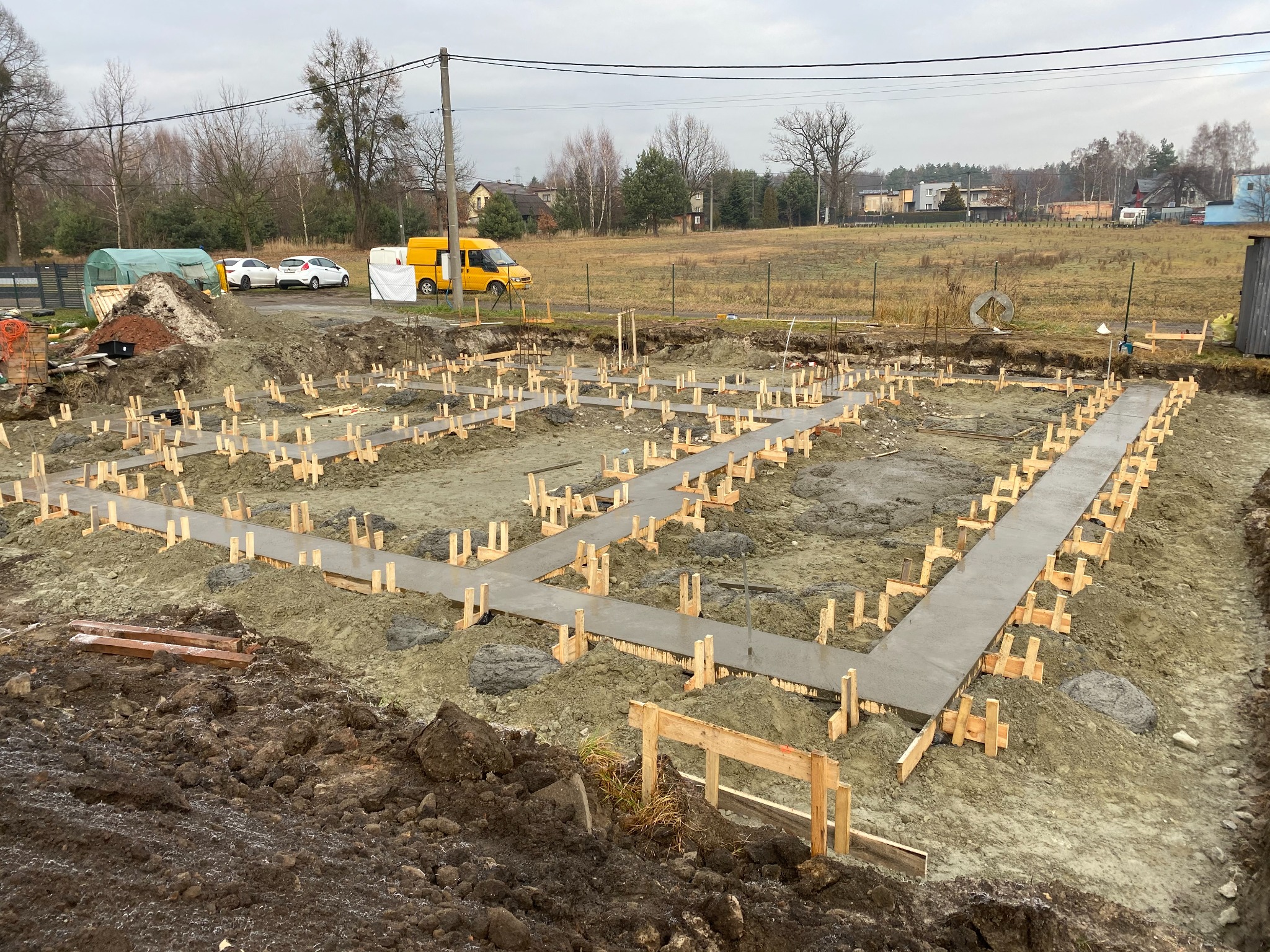 Wylane betonowe fundamenty pod budynek z drewnianym szalunkiem, widoczny teren budowy z żółtym busem w tle.
