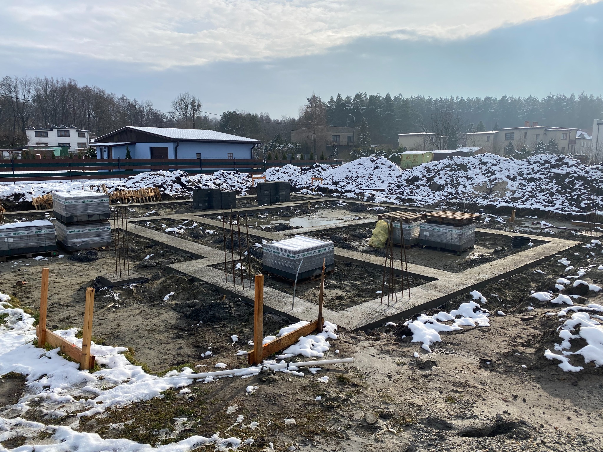Fundamenty domu jednorodzinnego w zimowej scenerii, z widocznymi zbrojeniami, bloczkami i drewnianymi szalunkami, częściowo pokryte śniegiem, w tle zabudowa jednorodzinna i las.