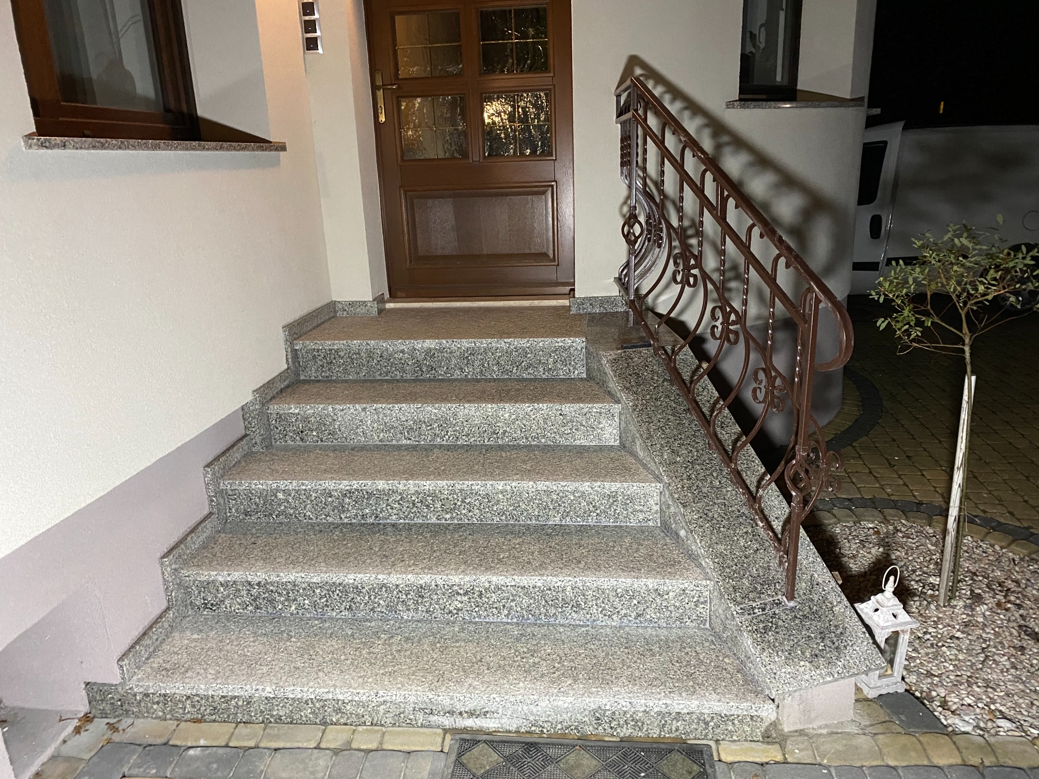 Granitowe schody zewnętrzne prowadzące do brązowych drzwi wejściowych, z kutą, ozdobną balustradą, oświetlone światłem zewnętrznym.