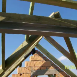 Konstrukcja drewniana dachu w trakcie budowy, widoczne krokwie, murłaty i fragment murowanej ściany szczytowej z cegły ceramicznej na tle błękitnego nieba. Na cegłach widoczny czarny element izolacji.