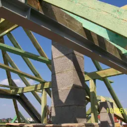 Konstrukcja drewniana dachu z elementami stalowymi i kominem z bloczków betonowych na tle błękitnego nieba, widoczne datowanie na zdjęciu.