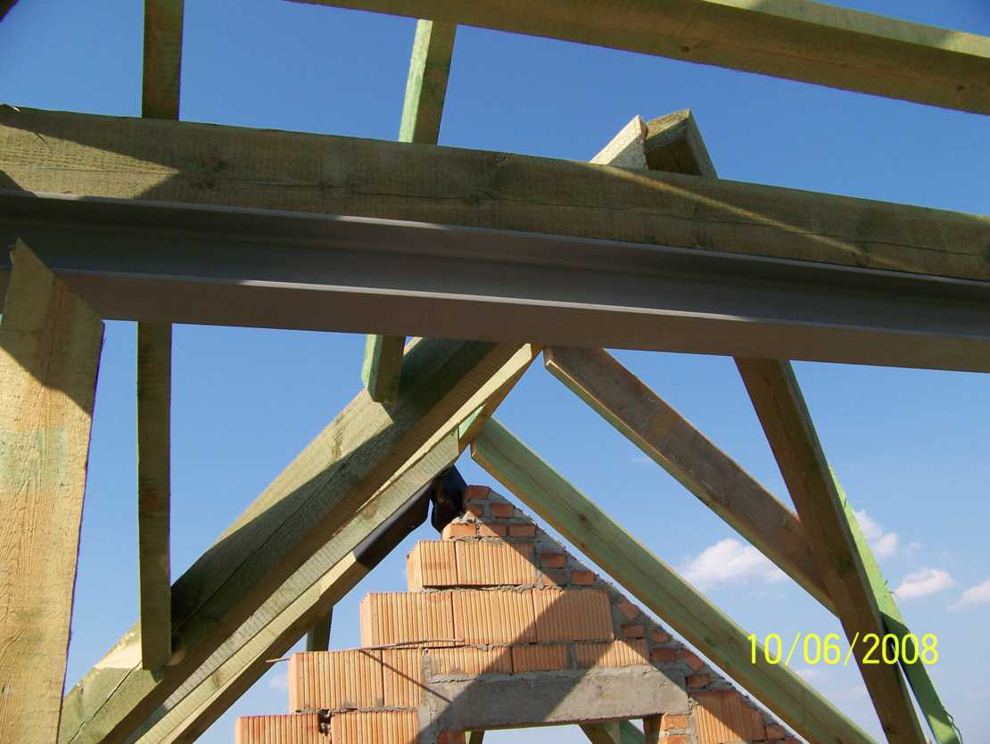 Konstrukcja drewniana dachu w trakcie budowy, widoczne krokwie, murłaty i fragment murowanej ściany szczytowej z cegły ceramicznej na tle błękitnego nieba. Na cegłach widoczny czarny element izolacji.