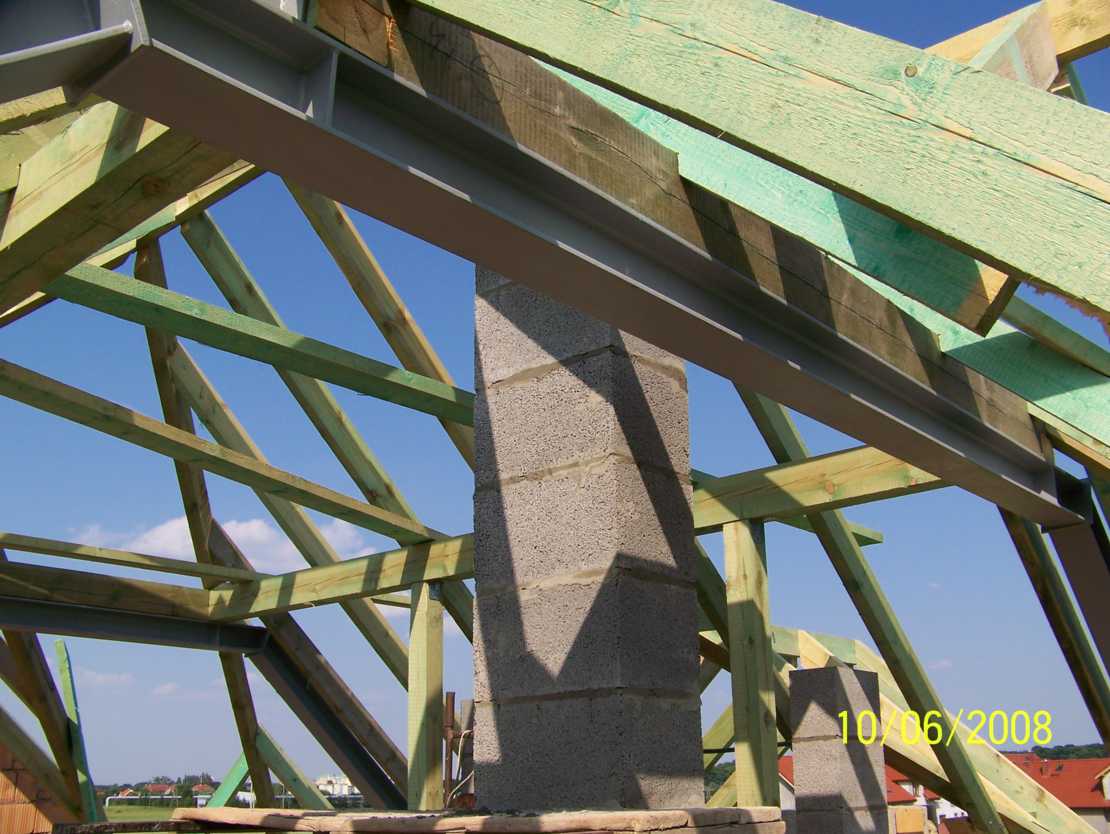 Konstrukcja drewniana dachu z elementami stalowymi i kominem z bloczków betonowych na tle błękitnego nieba, widoczne datowanie na zdjęciu.