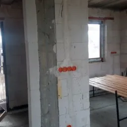 Surowy stan budynku w trakcie prac: widoczne puszki elektryczne w ścianie z betonu komórkowego, częściowo otynkowana ściana, okno w tle.