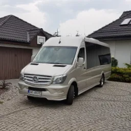 Beżowy bus Mercedes-Benz Sprinter na brukowanym podjeździe przed domem z brązową dachówką i panelami solarnymi.