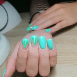 Dłonie z turkusowym manicure, kwadratowe paznokcie z efektem 'slime' w jaśniejszym odcieniu zieleni, lampa UV w tle.