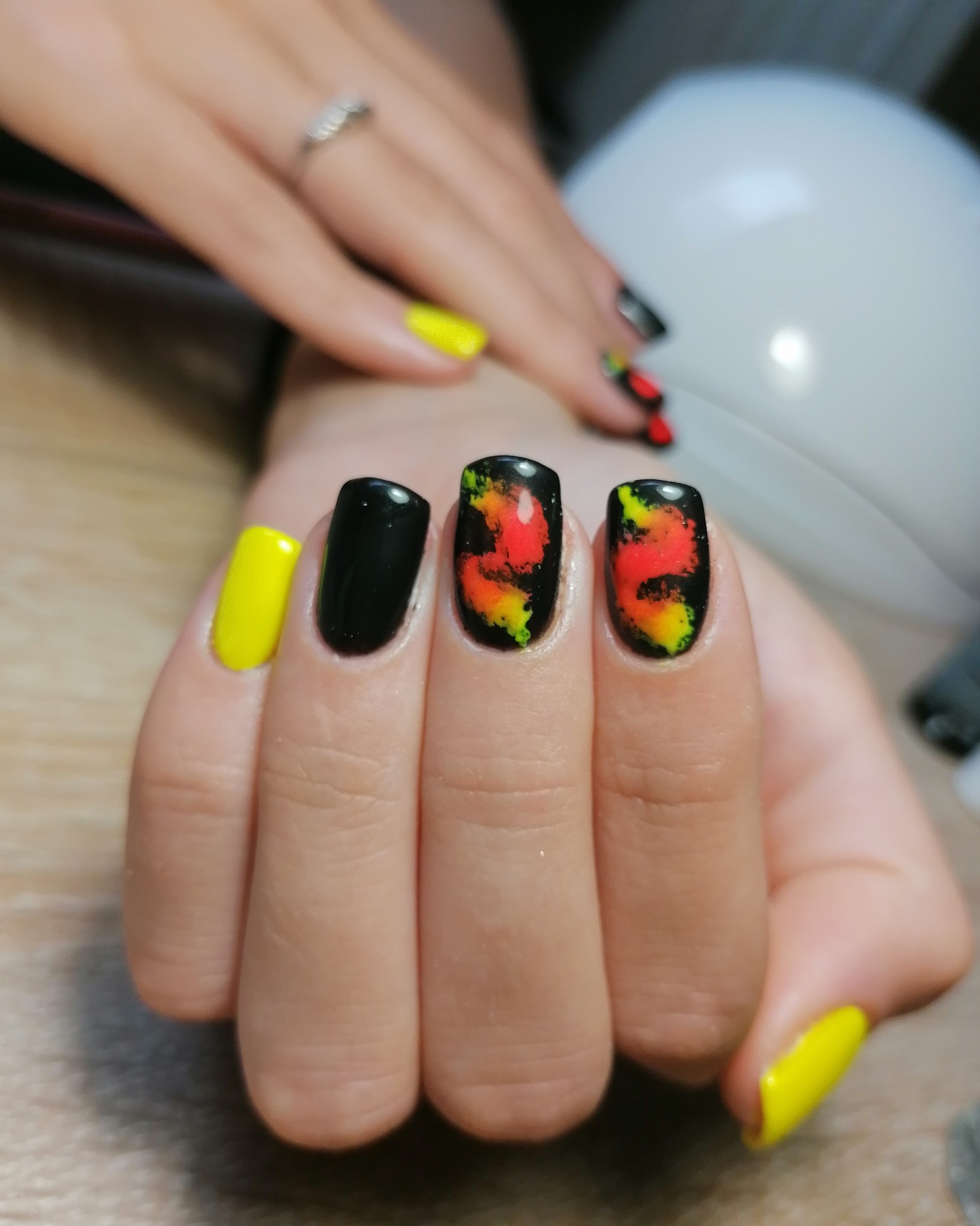 Dłoń z pomalowanymi paznokciami: dwa żółte, jeden czarny i dwa z czarnym tłem i abstrakcyjnym, ognistym wzorem w kolorach czerwieni, pomarańczy i zieleni.