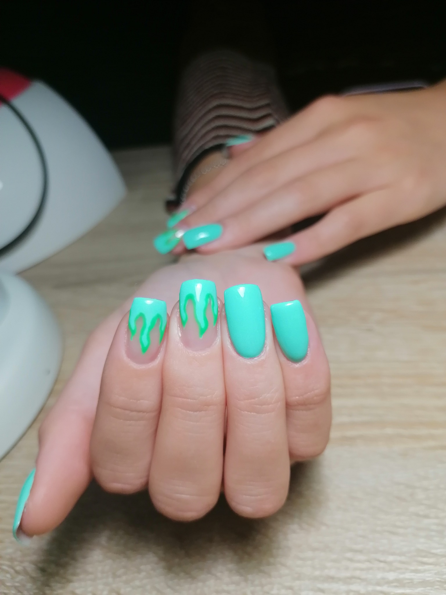 Dłonie z turkusowym manicure, kwadratowe paznokcie z efektem 'slime' w jaśniejszym odcieniu zieleni, lampa UV w tle.
