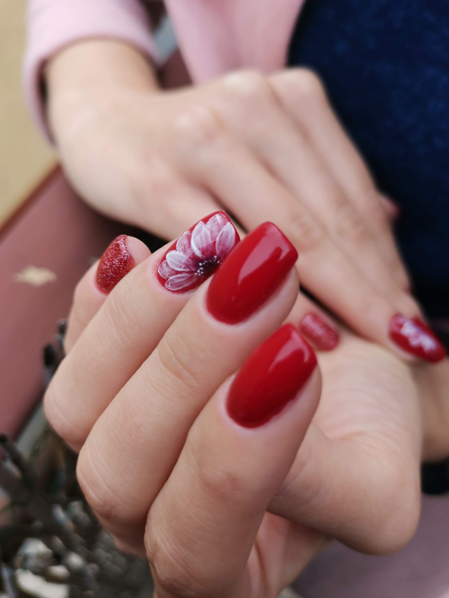 Czerwony manicure hybrydowy z brokatowym akcentem i ręcznie malowanym białym kwiatem na jednym paznokciu.