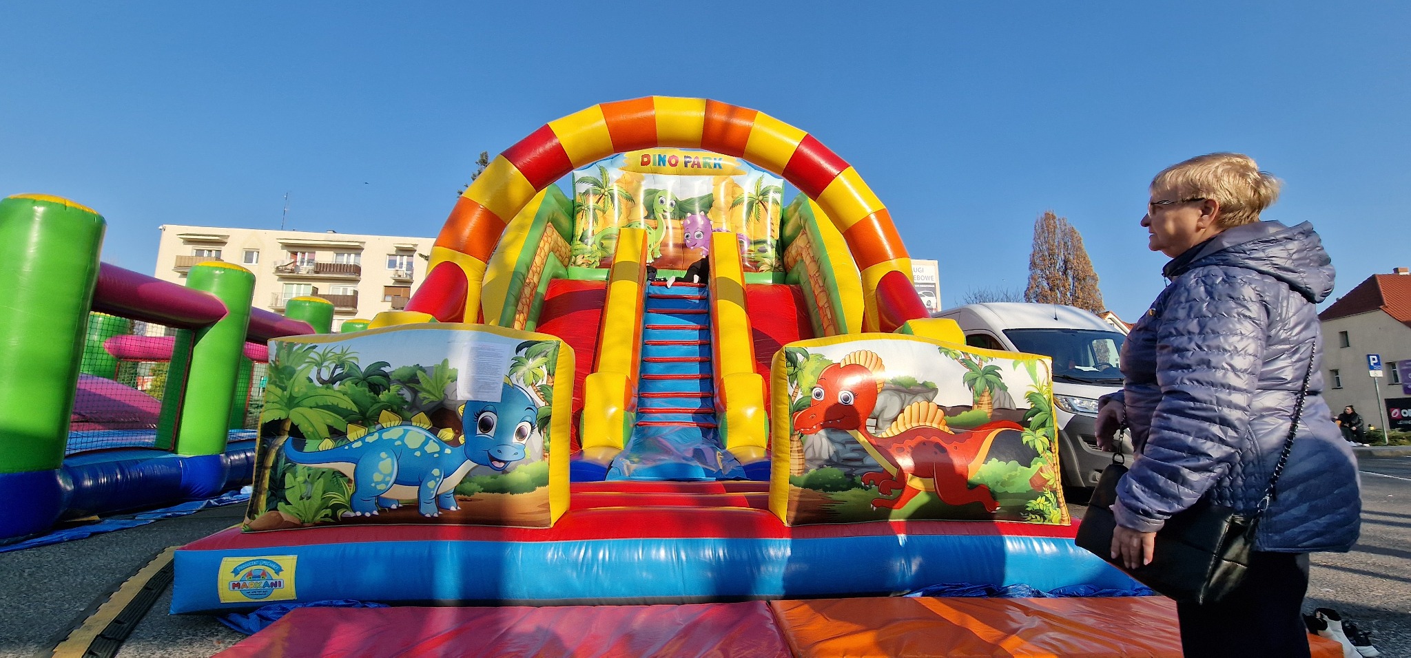 Kolorowy dmuchany zamek z motywem dinozaurów, widoczna kobieta w kurtce, budynki w tle.
