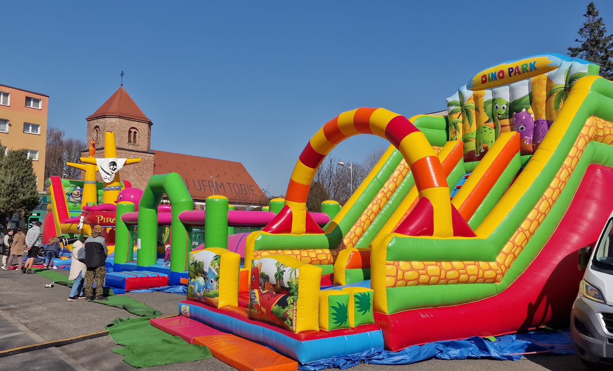 Kolorowy, dmuchany plac zabaw z motywem dinozaurów i piratów, ustawiony na zewnątrz, z widocznymi elementami architektonicznymi w tle oraz bawiącymi się dziećmi.