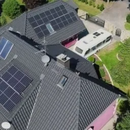 Widok z góry na dom z ciemnoszarą dachówką, na której zamontowane są panele słoneczne; na podjeździe zaparkowany biały bus.