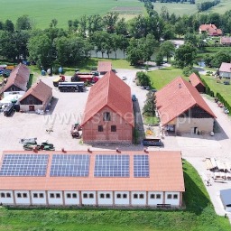 Widok z lotu ptaka na budynek gospodarczy z panelami słonecznymi na dachu, otoczony zielonym terenem i innymi budynkami.