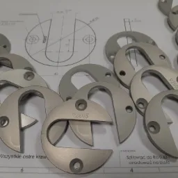 Partie anodowanych na biało, szlifowanych metalowych elementów w kształcie podkowy z otworem, oznaczonych numerem 92605, leżą na rysunku technicznym z wymiarami.