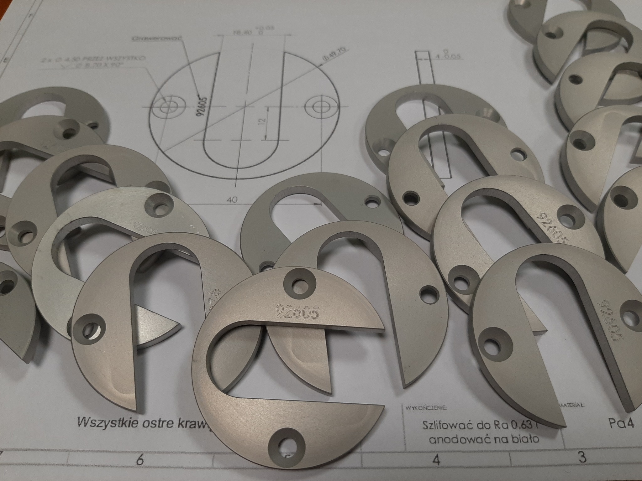 Partie anodowanych na biało, szlifowanych metalowych elementów w kształcie podkowy z otworem, oznaczonych numerem 92605, leżą na rysunku technicznym z wymiarami.