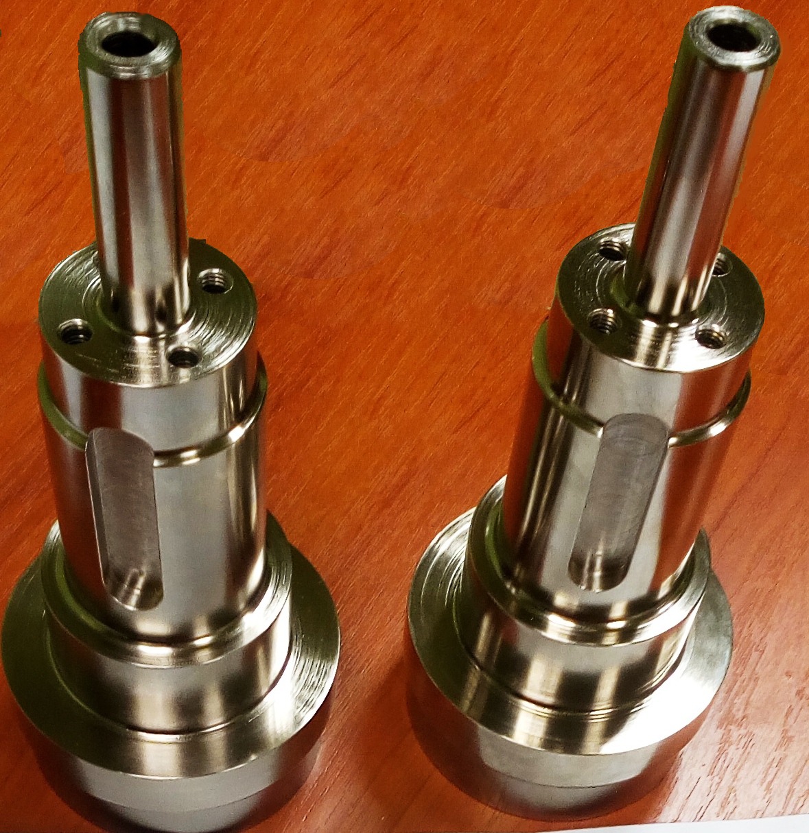 Dwa identyczne, precyzyjnie wykonane, metalowe elementy z otworem, leżące na drewnianej powierzchni. Widoczne ślady obróbki skrawaniem.