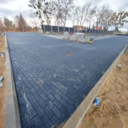 Ułożona, ciemnoszara kostka brukowa na dużej powierzchni placu, z widocznym paletami kostki, ubijarką wibracyjną i krawężnikiem, otoczona piaskiem i ziemią.