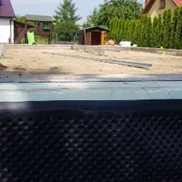 Fundament domu szkieletowego z izolacją przeciwwilgociową, widoczne elementy konstrukcyjne i przygotowany teren budowy w słoneczny dzień.