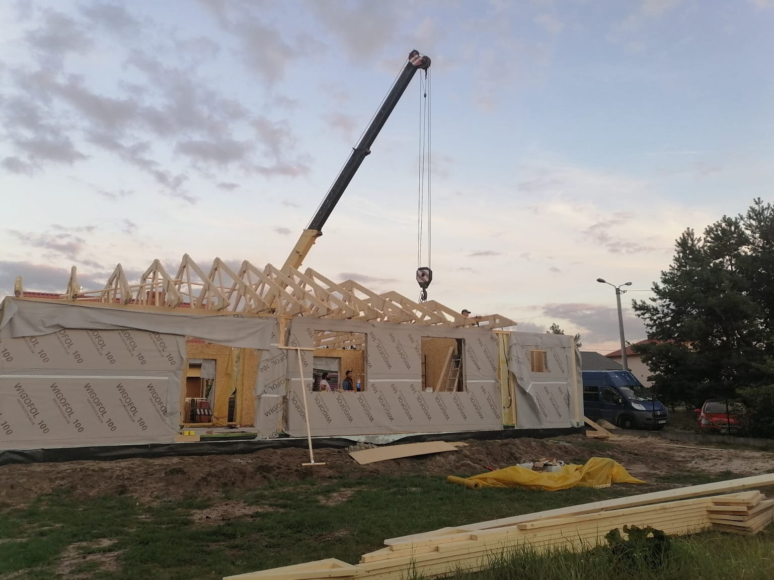 Montaż drewnianej konstrukcji dachu domu szkieletowego z użyciem dźwigu, widoczne ściany z izolacją Wigopol 100, w tle zaparkowane busy