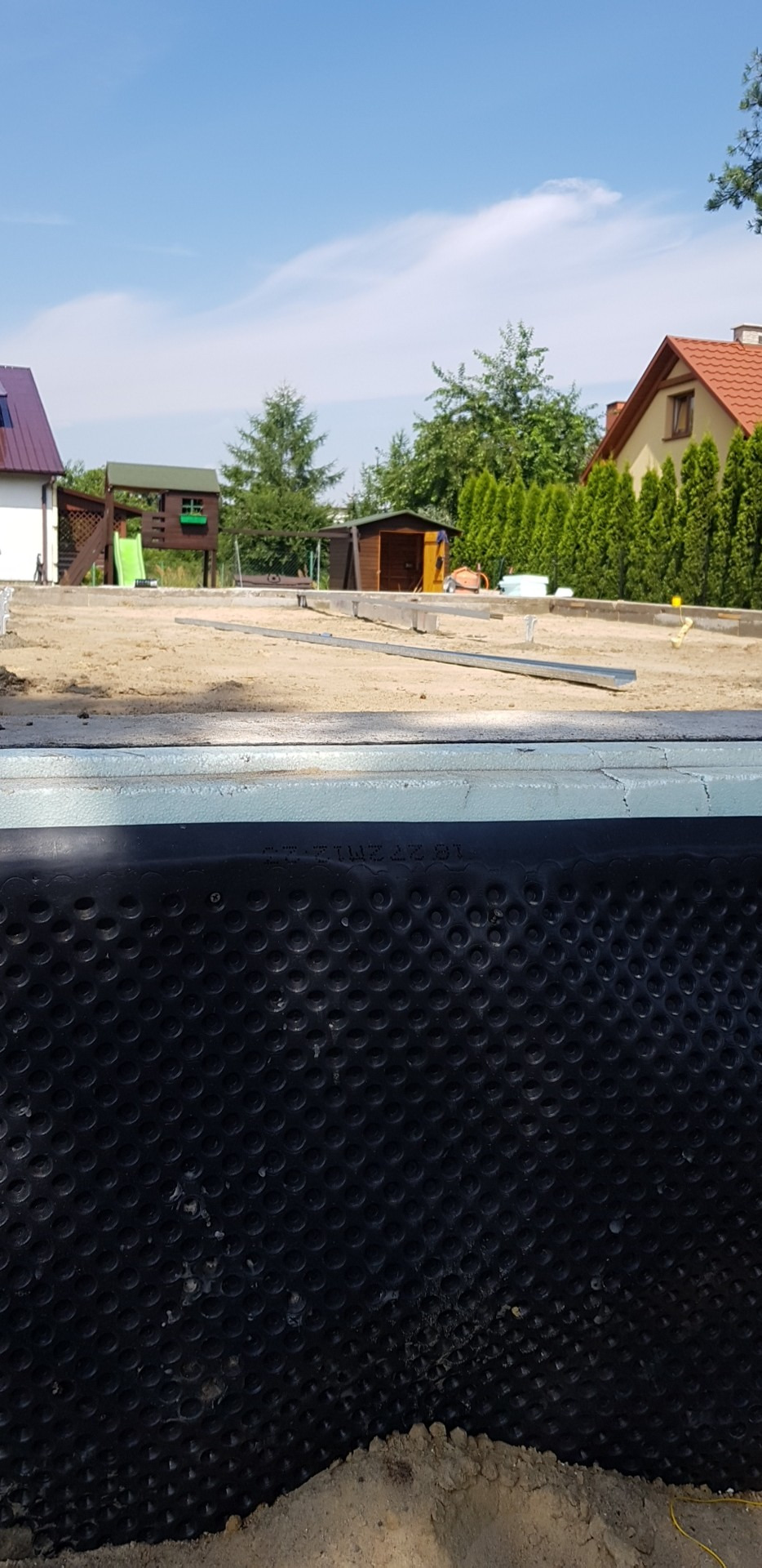 Fundament domu szkieletowego z izolacją przeciwwilgociową, widoczne elementy konstrukcyjne i przygotowany teren budowy w słoneczny dzień.