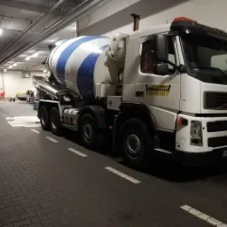 Betonomieszarka Volvo z biało-niebieskim bębnem, zaparkowana w hali garażowej oświetlonej lampami jarzeniowymi, widoczne logo firmy P.W. Adam na kabinie.