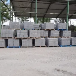 Stosy szarych płyt drogowych ułożonych na drewnianych i niebieskich plastikowych paletach pod zadaszeniem magazynu, z widoczną pryzmą piasku w tle.