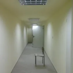 Korytarz biurowy z szarą podłogą z płytek, świeżo pomalowanymi kremowymi ścianami, oświetleniem sufitowym i krzesłem na środku. Widok w głąb korytarza.