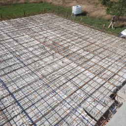 Zbrojenie płyty fundamentowej z siatki stalowej przed zalaniem betonem, widoczny pracownik i samochód dostawczy w tle.