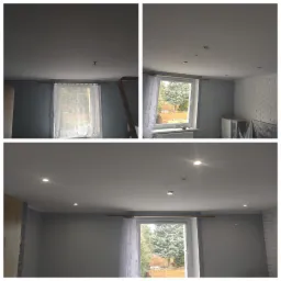 Pokój po montażu oświetlenia sufitowego, widoczne trzy punkty świetlne w suficie, ściany w odcieniach szarości, okno z firanką, fragment drabiny malarskiej i pozostałości po instalacji.