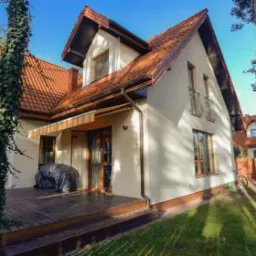 Gotowy dom jednorodzinny z jasną elewacją, czerwoną dachówką i drewnianym tarasem, otoczony zielenią i drzewami iglastymi.