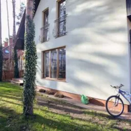 Jasny, dwukondygnacyjny dom jednorodzinny z drewnianymi oknami i kutymi balkonami, otoczony zielonym trawnikiem i drzewami, z rowerem opartym o ścianę przy wejściu.