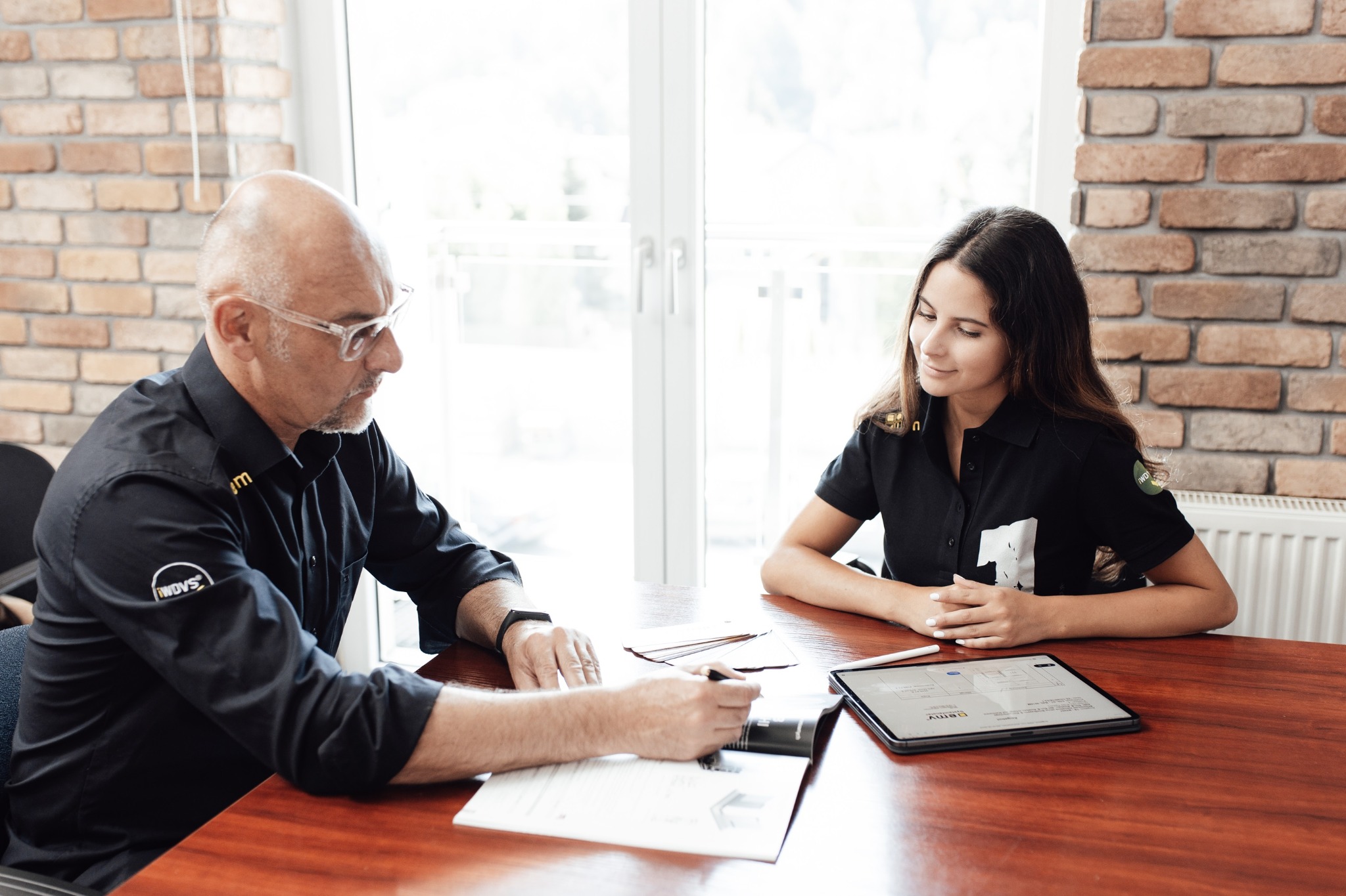 Spotkanie dwóch osób przy drewnianym stole: mężczyzna w koszuli z logo firmy wypełnia dokumenty, obok kobieta z tabletem, w tle ceglana ściana i okno.