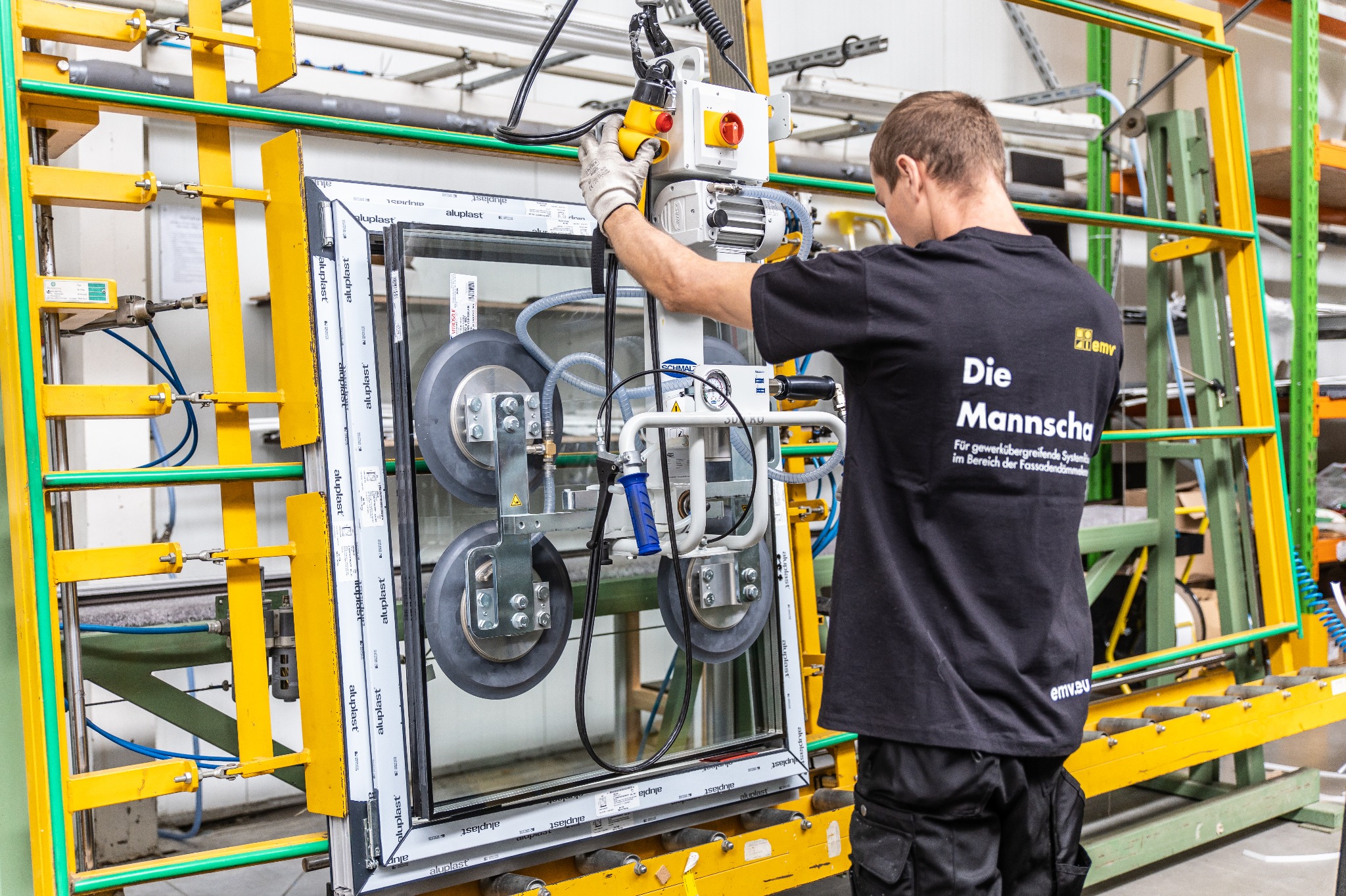 Pracownik w rękawicach obsługuje maszynę z przyssawkami, podnoszącą ramę okienną z szybą w hali produkcyjnej. Widoczny fragment żółto-zielonej konstrukcji wsporczej i napis na koszulce pracownika.