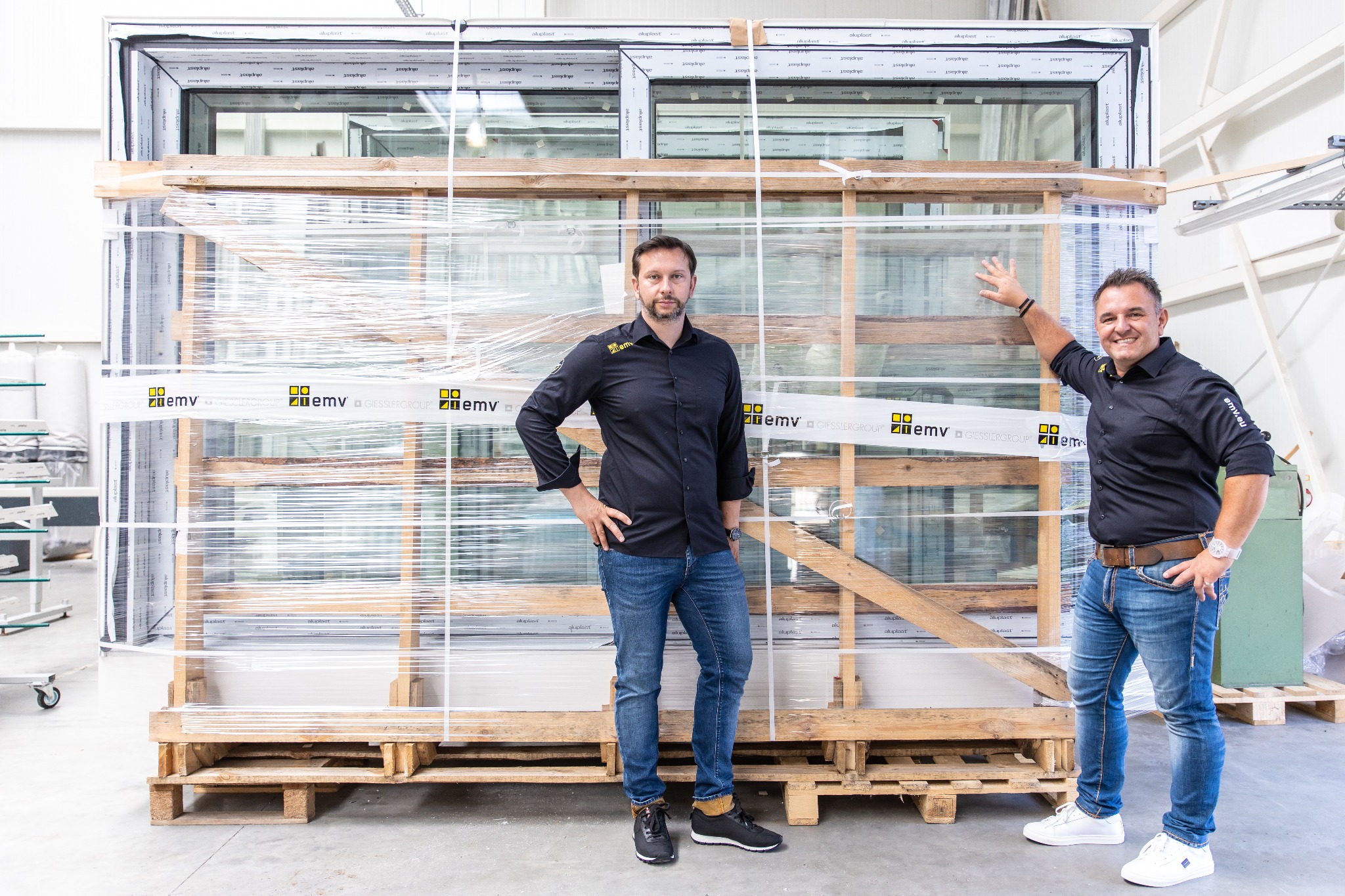 Dwaj mężczyźni prezentują zapakowane okna na palecie, widoczne logo producenta na folii ochronnej, obaj ubrani w koszule z logiem firmy.