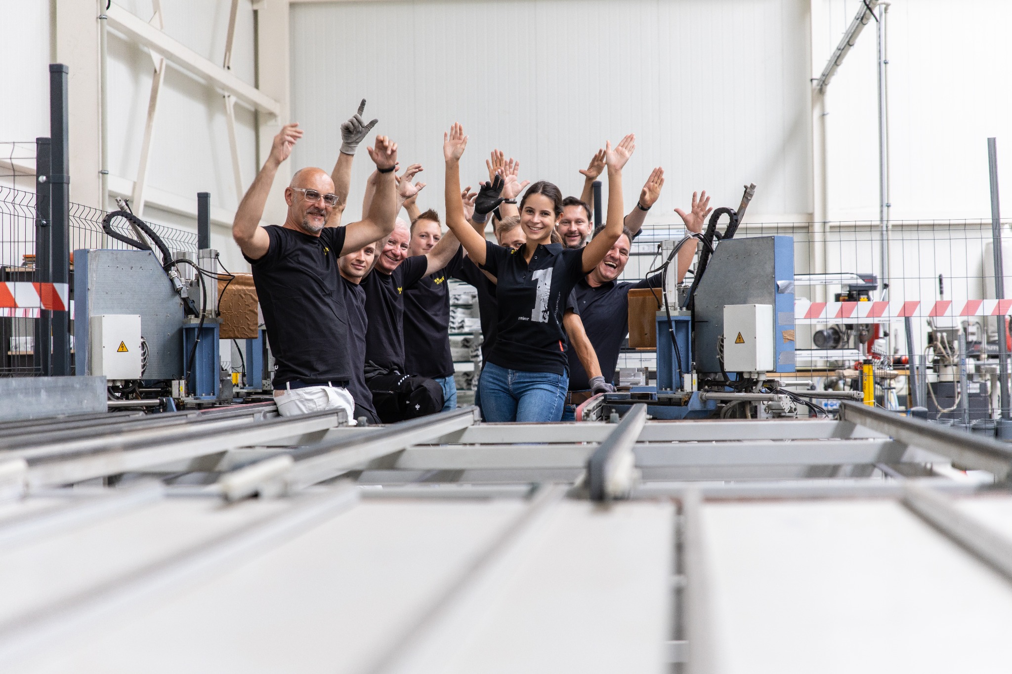 Grupa uśmiechniętych pracowników fabryki okien i drzwi PCV i aluminiowych, wznosząca ręce w geście radości na tle linii produkcyjnej z profilami aluminiowymi.