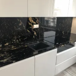 Czarny blat z kamienia z wyraźnym złotym użyleniem, wkomponowany w białą, lakierowaną zabudowę kuchenną, z widocznym zlewem i płytą indukcyjną.
