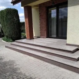 Zewnętrzne, trzybiegowe schody z brązowego kamienia prowadzące do domu z brązowymi drzwiami przesuwnymi i kolumnami z czerwonej cegły, otoczone kostką brukową i zielenią.